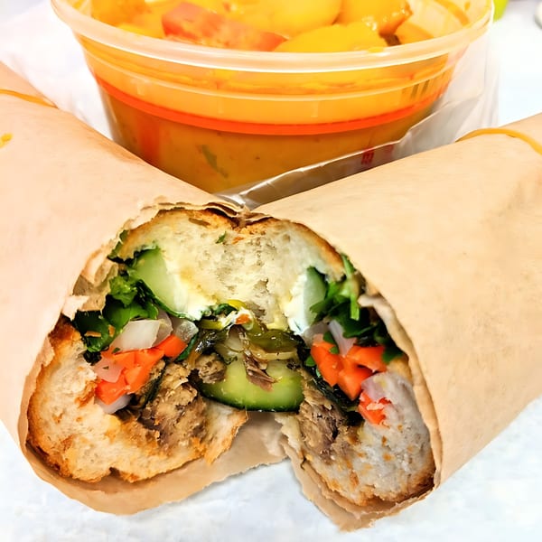 A Vietnamese banh mi sandwich, cut in half, with a side of soup in a plastic container.