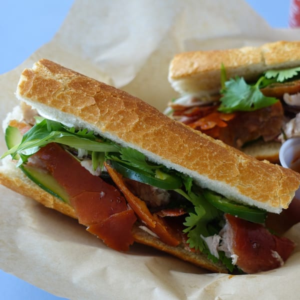 Two O Banh Mi sandwiches with crispy pork, cilantro, and jalapeños on a light brown paper.